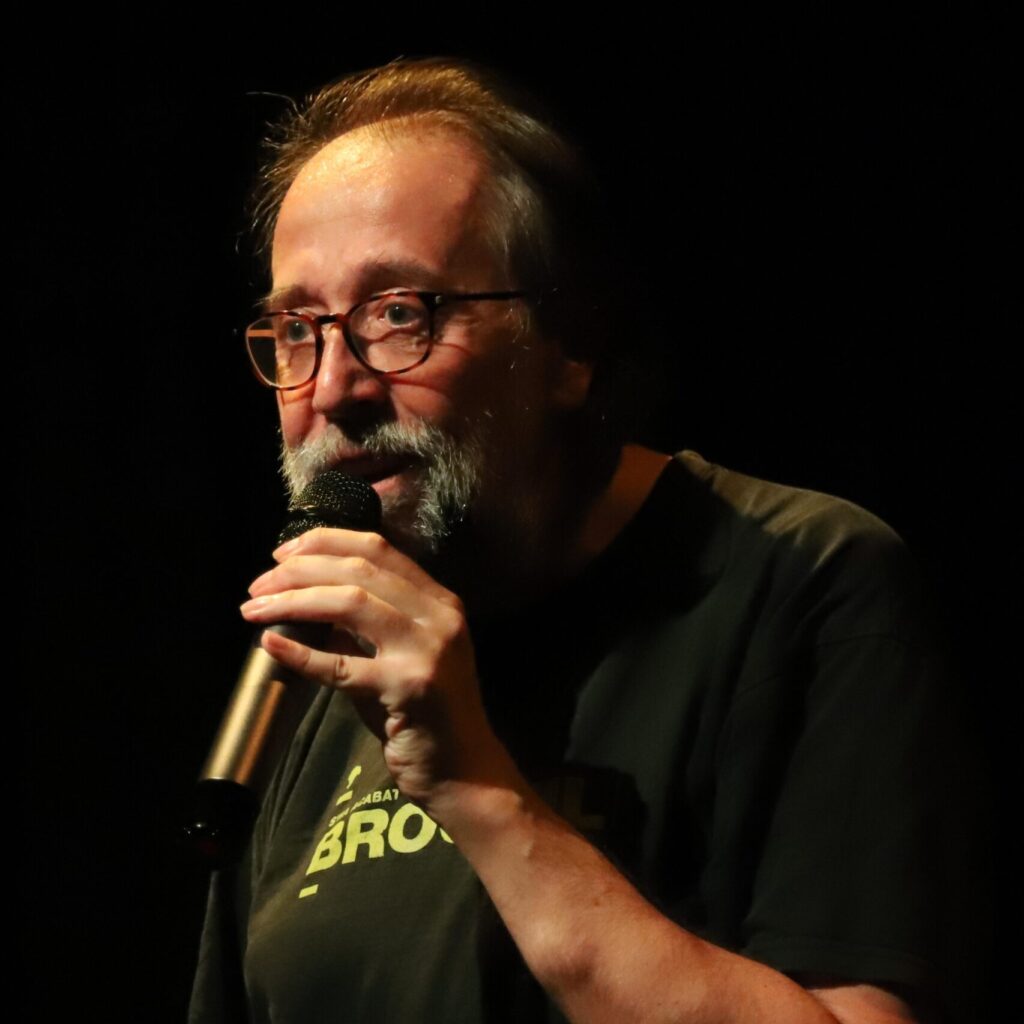 Comedian Chris Groves speaks into microphone in black shirt